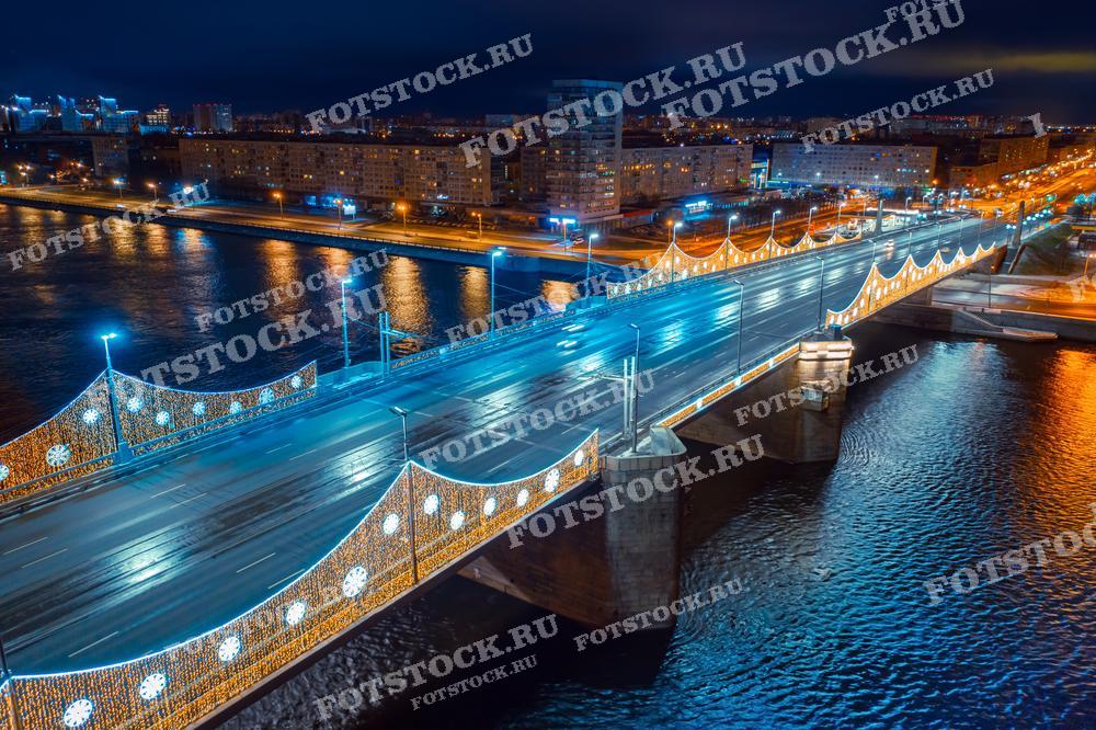 На фотографии изображен Володарский мост в Санкт-Петербурге ночью. Мост украшен праздничной иллюминацией, сверкающей в темноте. На заднем плане видна река Нева и городские огни, отражающиеся в воде. Зимний вечер добавляет атмосферу уюта и праздника.